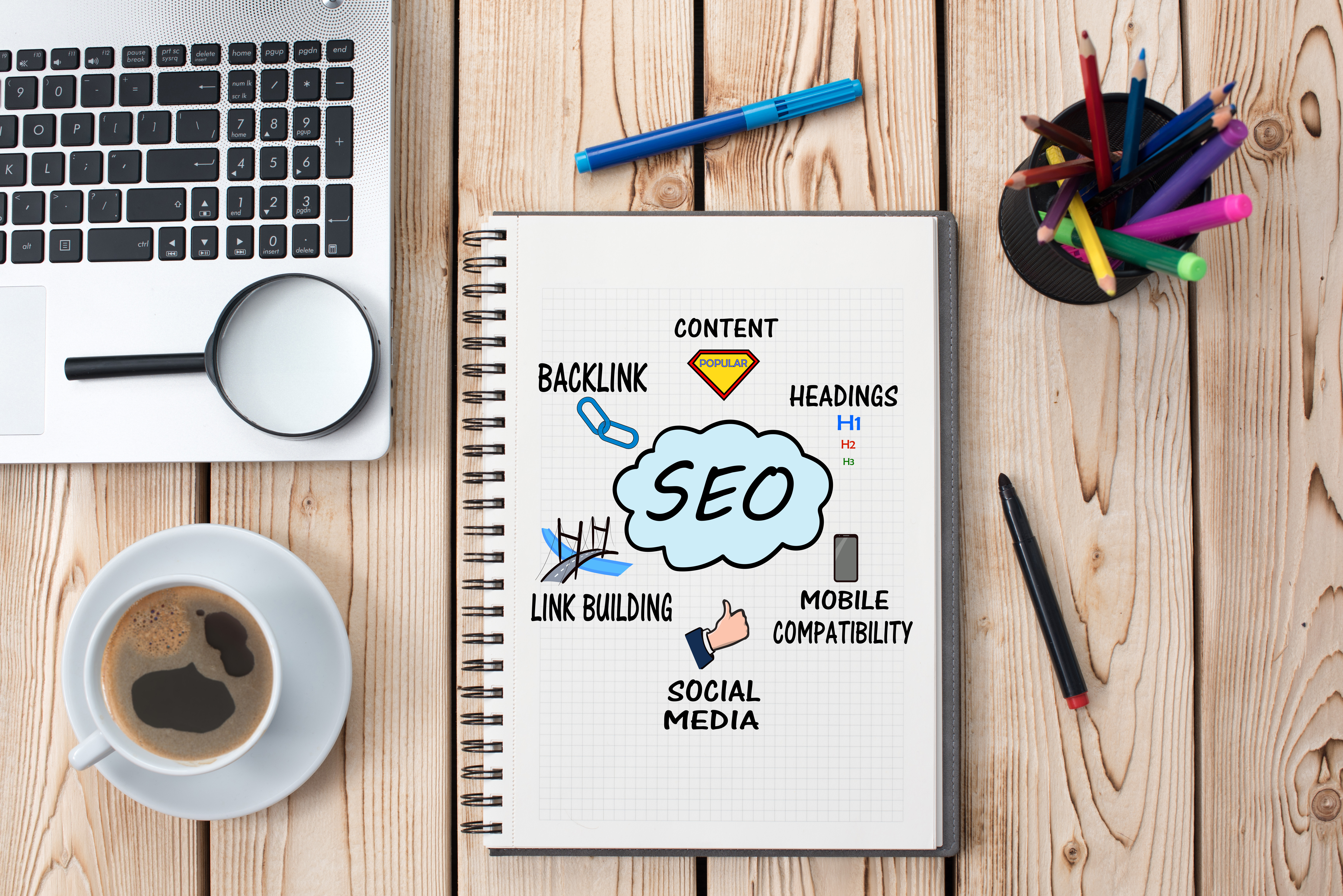 Desk with laptop, magnifying glass, pens and pencils, coffee, and notebook with Search Engine Optimization clipart.